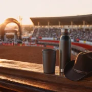 Copo, garrafa térmica e chapéu sertanejo diante de uma arena de rodeio ao pôr do sol