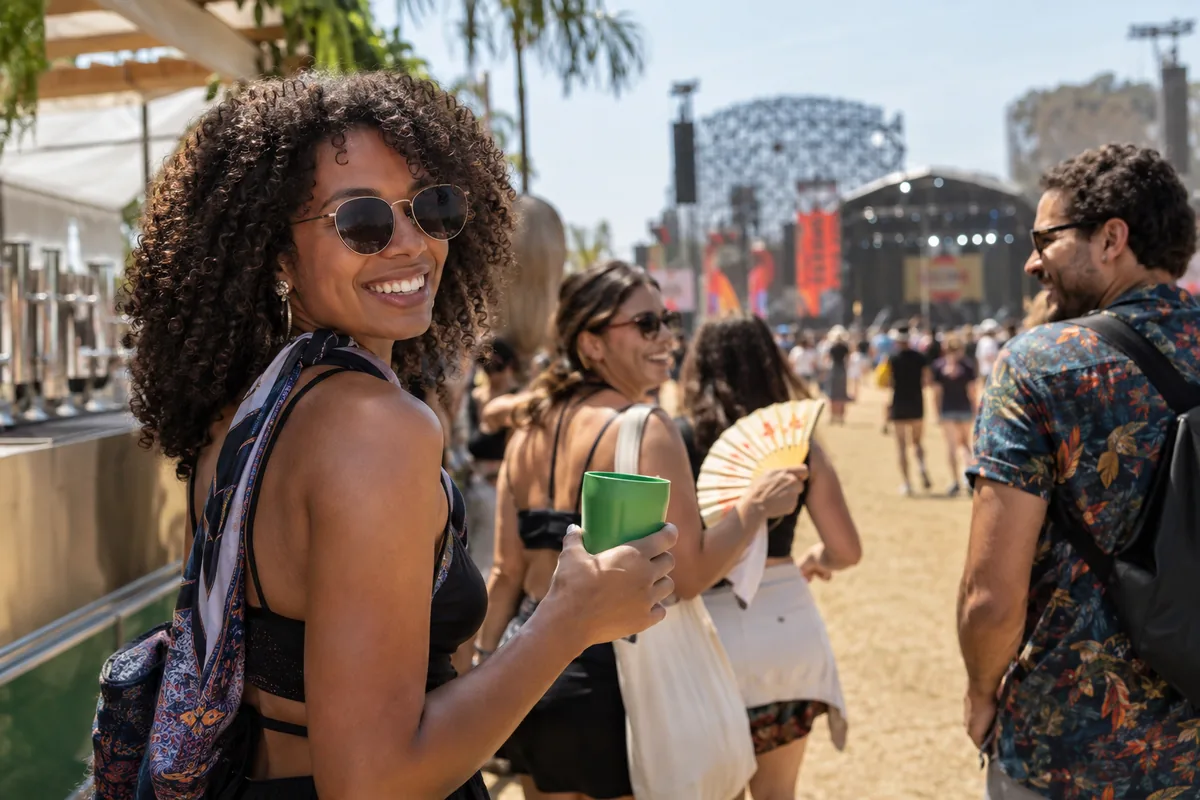 Participante de festival ao ar livre segura um copo enquanto outra pessoa usa um leque em um dia de sol.